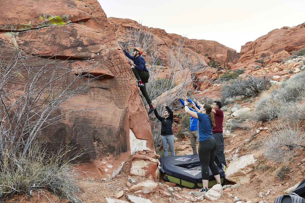 A climber going up a boulder with spotters beneath him standing around a crash pad.