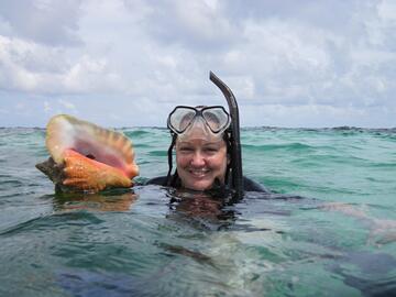 Carrie Tyler in the water wearing scuba gear and smiling while holding a large seashell above the surface 