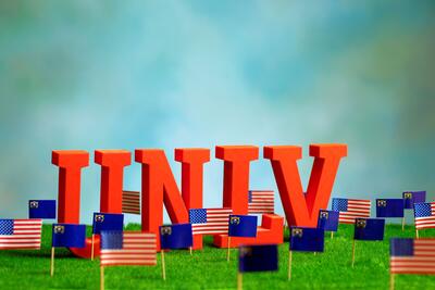 Red UNLV letters sitting on grass with US and Nevada flags around it.