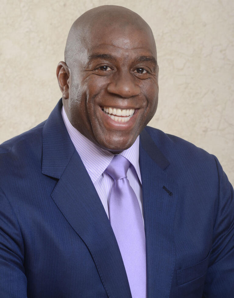 Portrait of Magic Johnson wearing a navy suit with a lavendar shirt and tie.