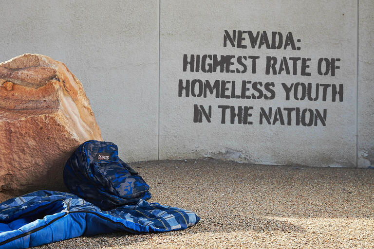 backpack and sleeping bag next to wall with &quot;Nevada: Highest Rate of Homeless Youth in the Nation&quot; spray painted on it