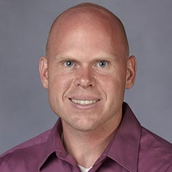 A smiling man with shaven hair wearing a purple collared shirt.