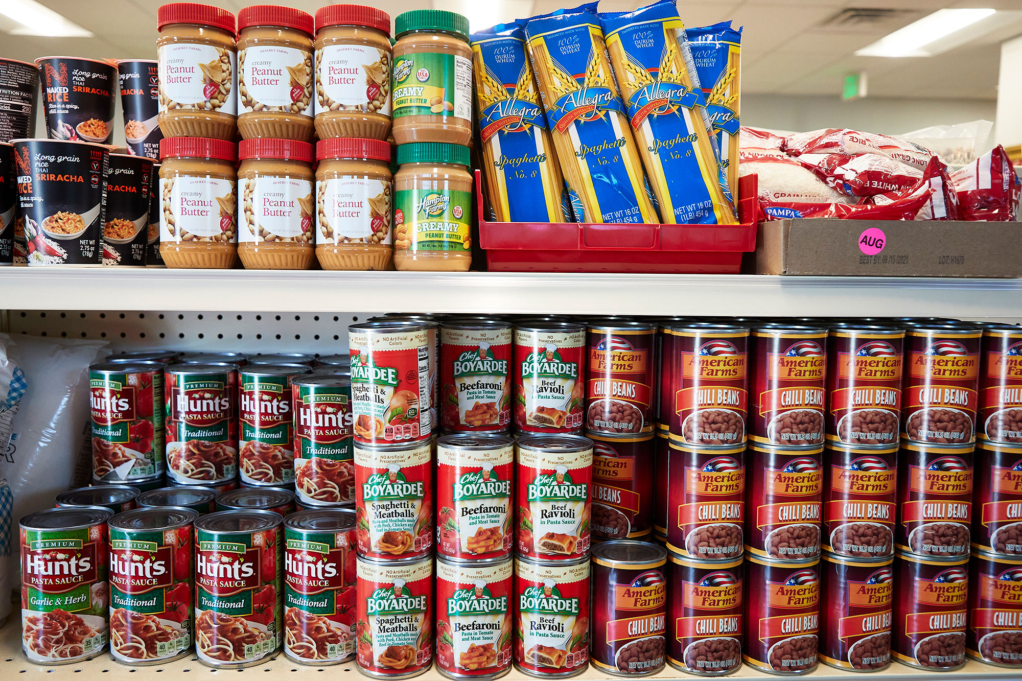 A shelf in a food pantry with various items including cans of pasta sauce, chili beans, Chef Boyardee meals, jars of peanut butter, and packs of spaghetti noodles.