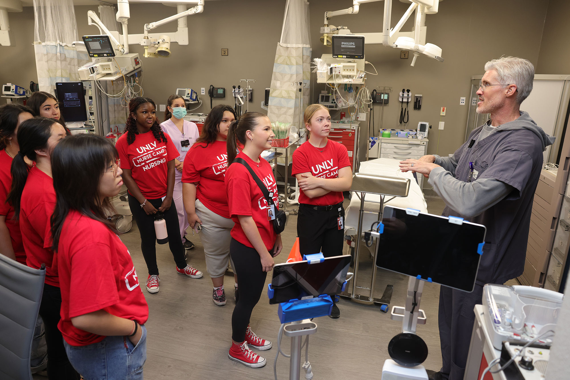 Students in red "UNLV Nurse Camp" shirts listen to an instructor in a clinical simulation lab with medical equipment and hospital beds.