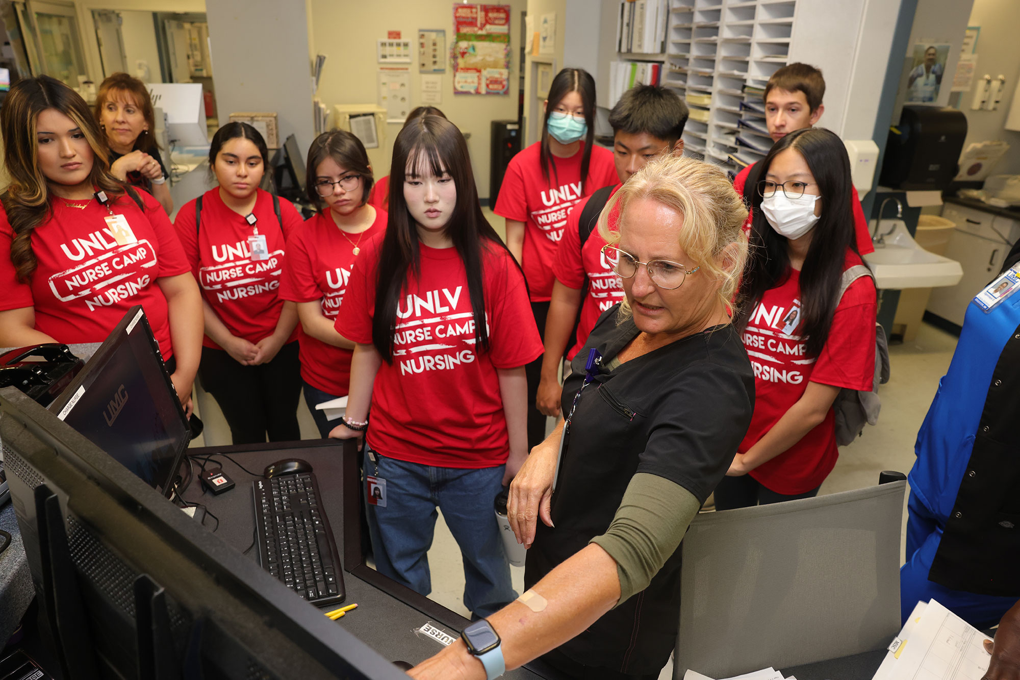 High school students wearing red "UNLV Nurse Camp Nursing" shirts gather around a healthcare professional in a clinical setting.