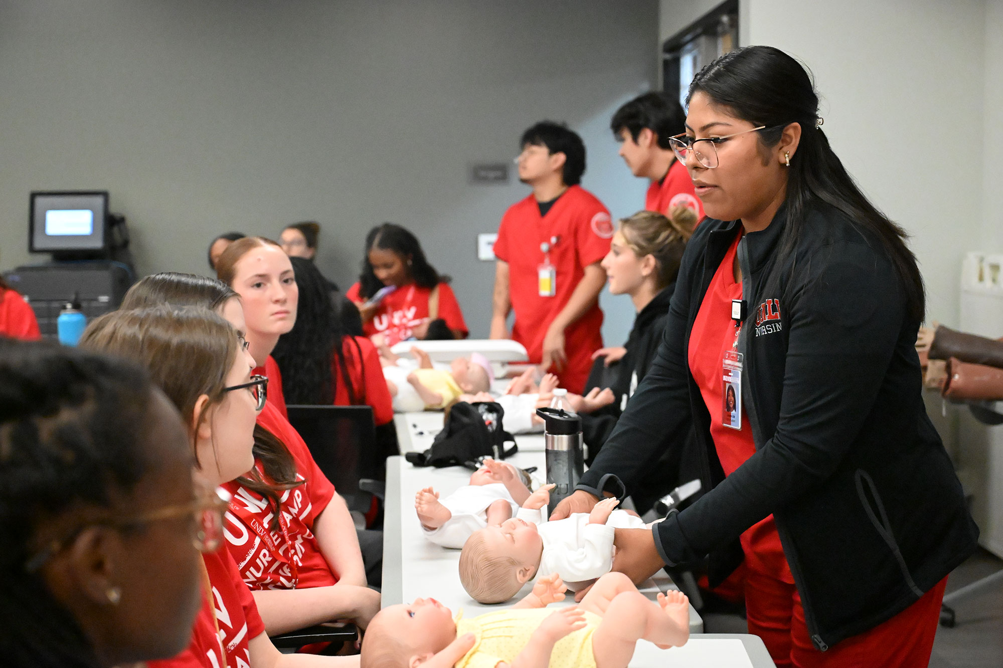 Nursing camp students observe an instructor demonstrating infant care using baby mannequins in a classroom.