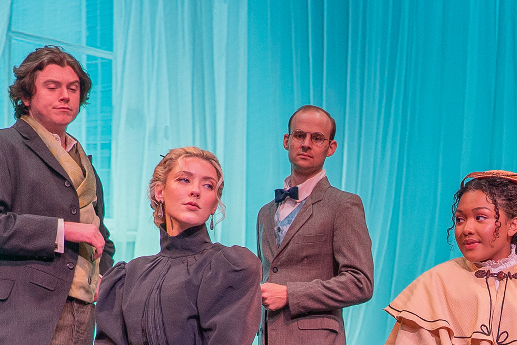 Two women in victorian attire sitting on a bench on a stage as two men standing behind them look down towards them