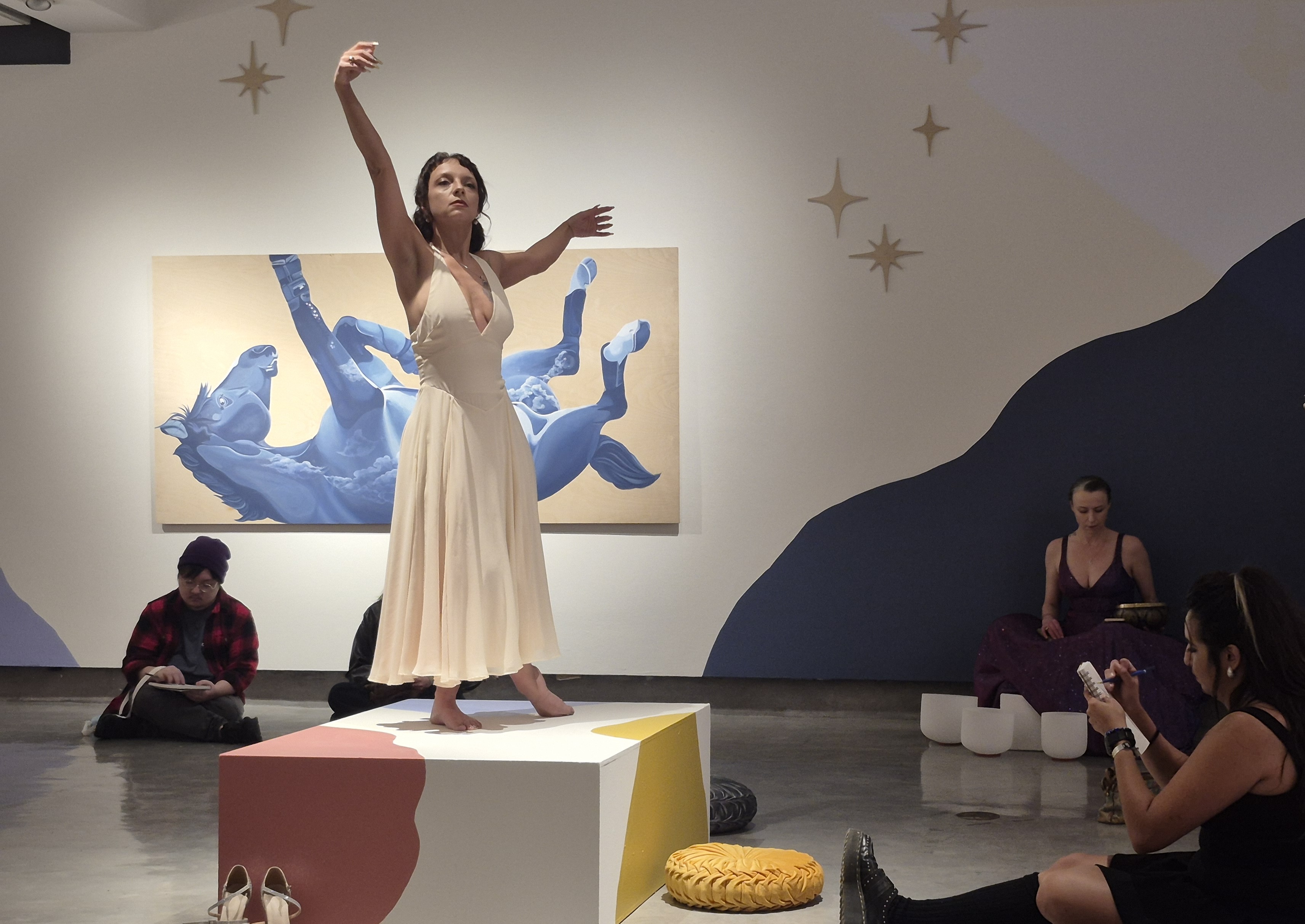 Woman in a white dress posing on a white box with a red, white, and brown blob painted onto the box with students sitting on the floor sketching the model
