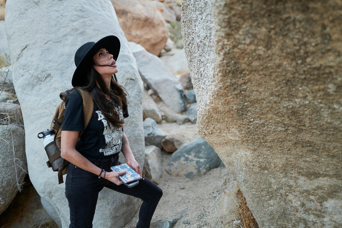 a woman stands in a stony desert landscape with a notebook in her hand, looking up at an immense rock.