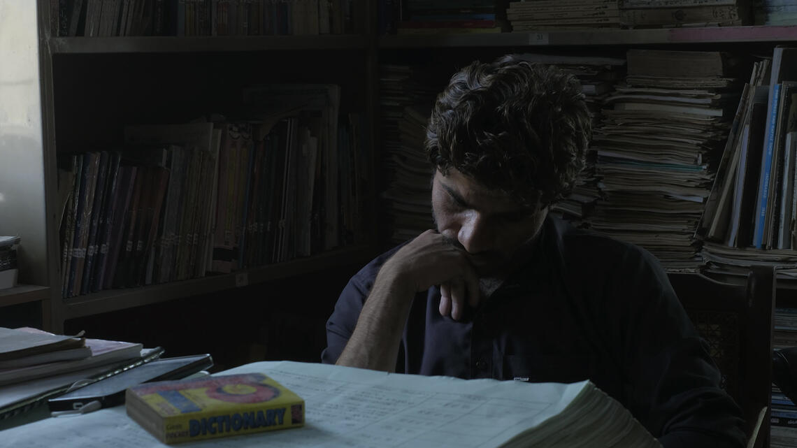 A man sits with his chin on his hand looking down in concentration at a heap of printed pages on the table in front of him. The room around him is lined with overflowing bookshelves.