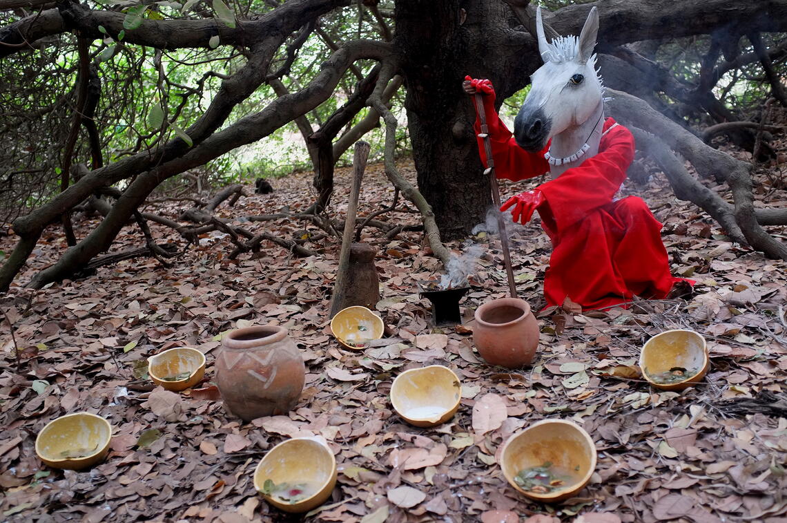 A red-robed figure wearing a white horse head mask sits under a tree. With one red-gloved hand it gestures to an arrangement of bowls and pots lying on the leaf litter. The other hand rests on a wooden cane. The bowls hold a soup of liquid and leaves.  Smoke rises from a black vessel by the figure’s side.