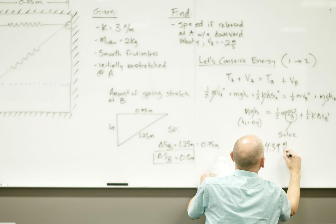 A professor writing equations on a blackboard with a marker.