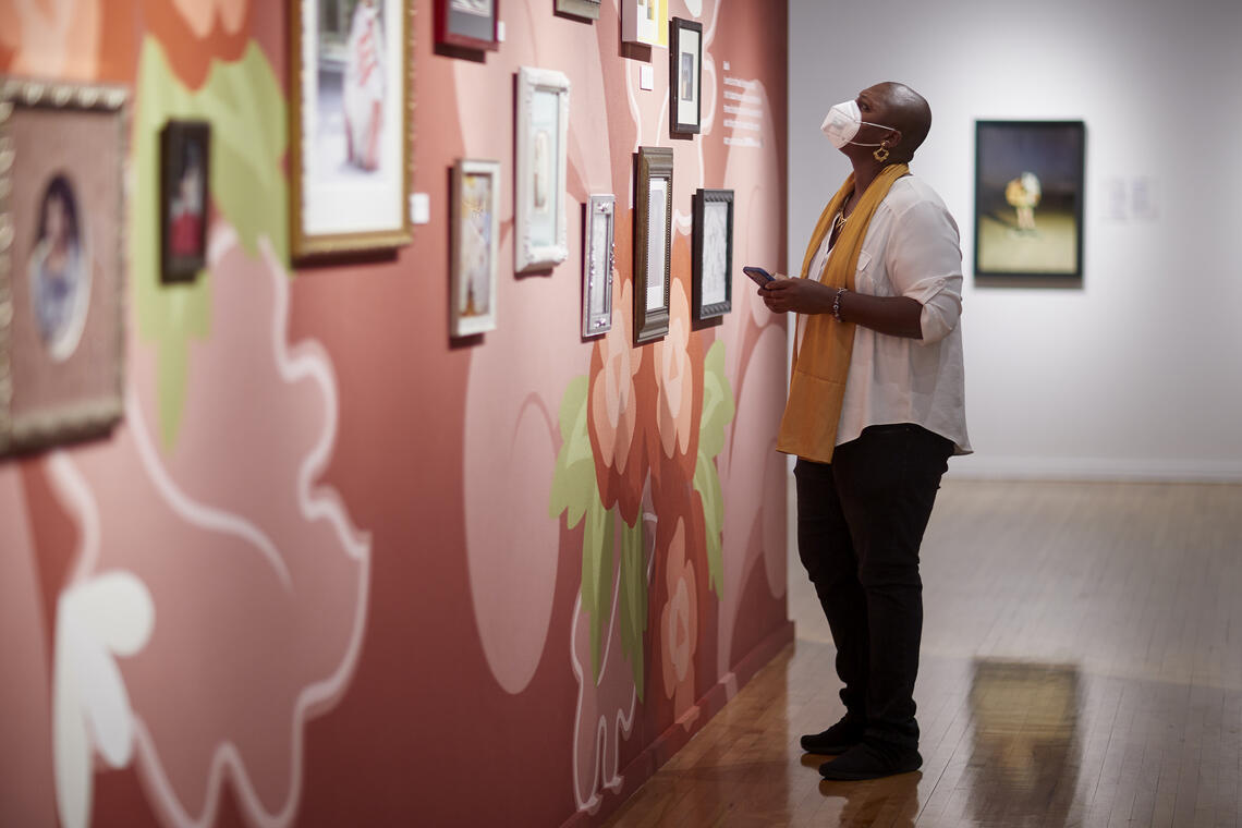 A museum visitor standing in profile looks at a wall with many portraits of black women on display in the exhibition Seeing/Seen. The portraits are hung on a wall that has been painted with a floral design.