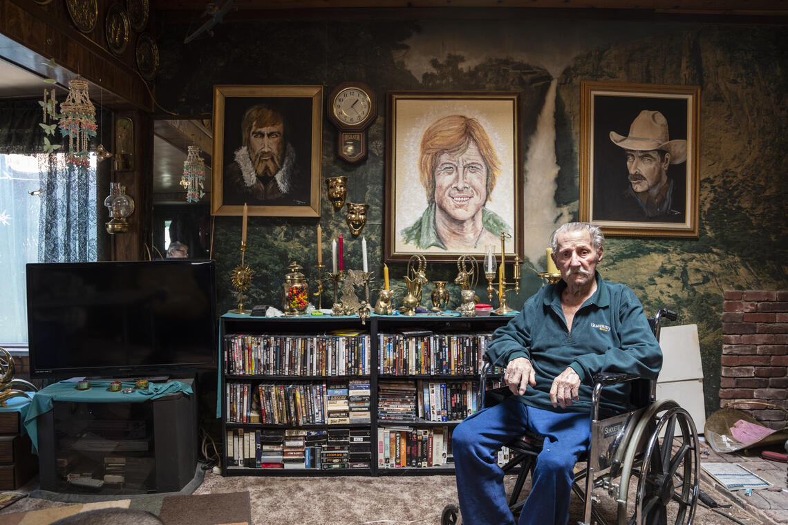 ARTIST MONTY BRANNIGAN, Darwin, CA, elderly sits in a wheel chair looking directly at the camera. There are paintings and books and ephemera behind him.