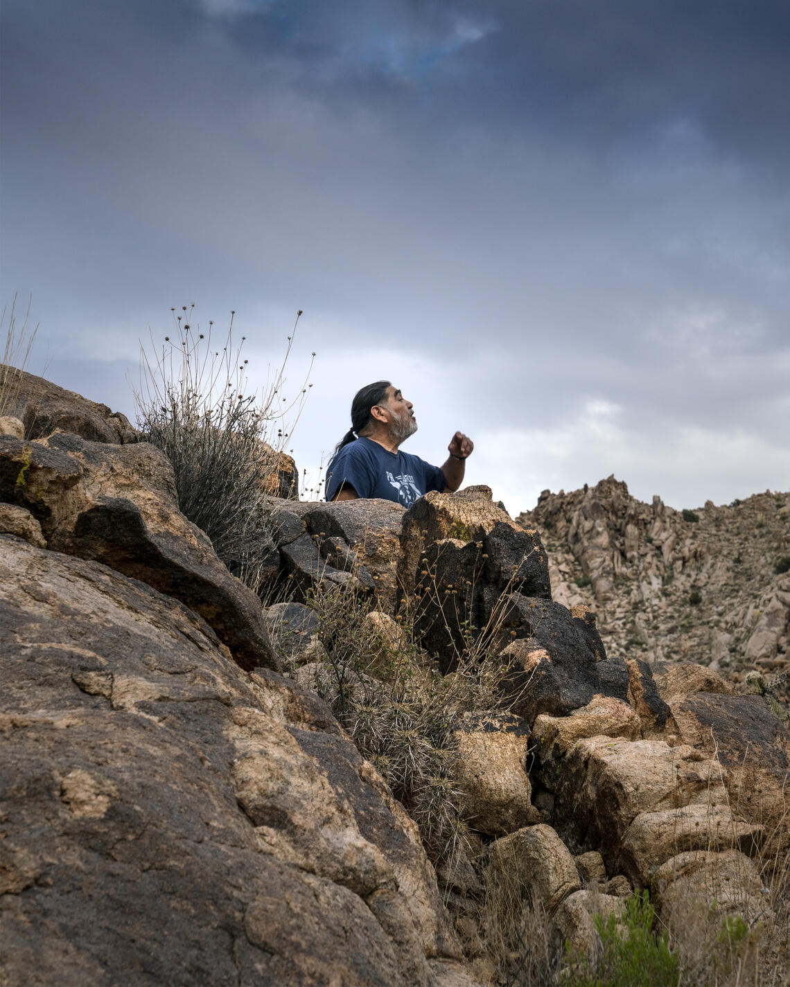 A native American man in a blue t-shirt sits atop a rocky desert outcrop and sings. A cloudy blue sky is behind him.