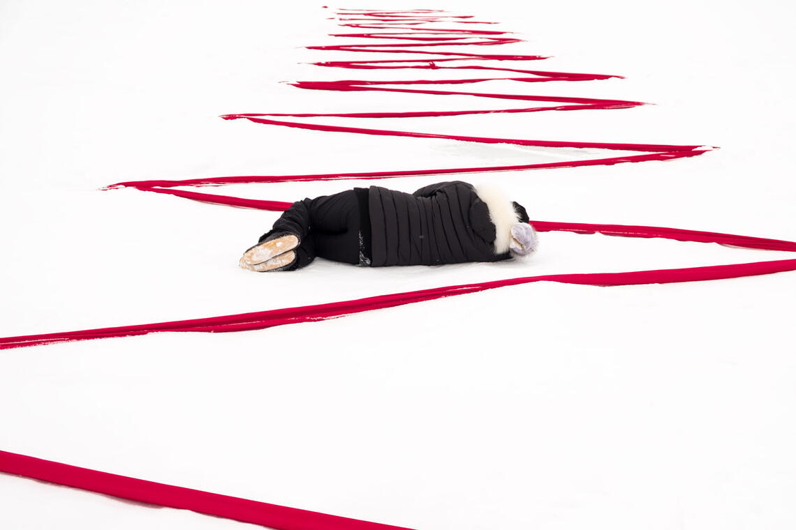 A figure in dark clothing lays on their side facing away from the viewer. They are laying in a snow white landscape, and bright red fabric zigzags across the snow and around the figure's body.