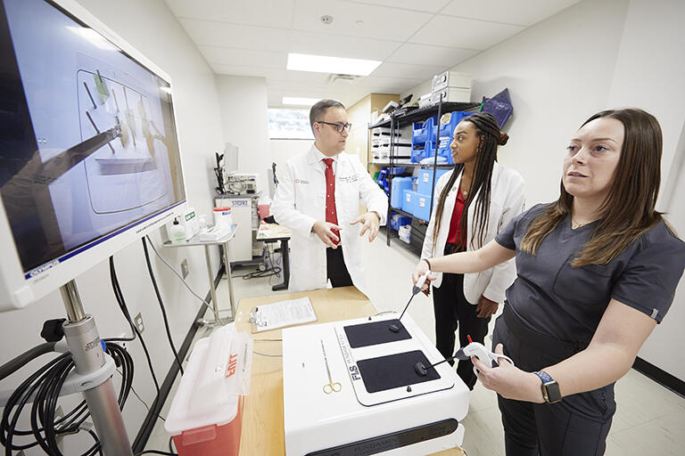 Woman working with a surgical practice tool and two others supervising her work