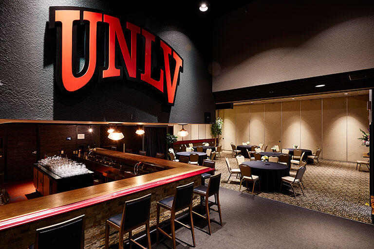 A vacant dining area with tables and chairs and a bar counter with barstools and UNLVs branding on the wall.
