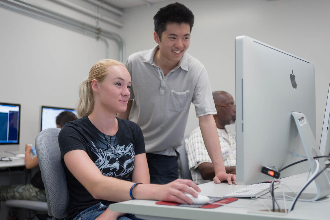 A woman sitting down at a computer with a man standing beside her looking at the monitor.