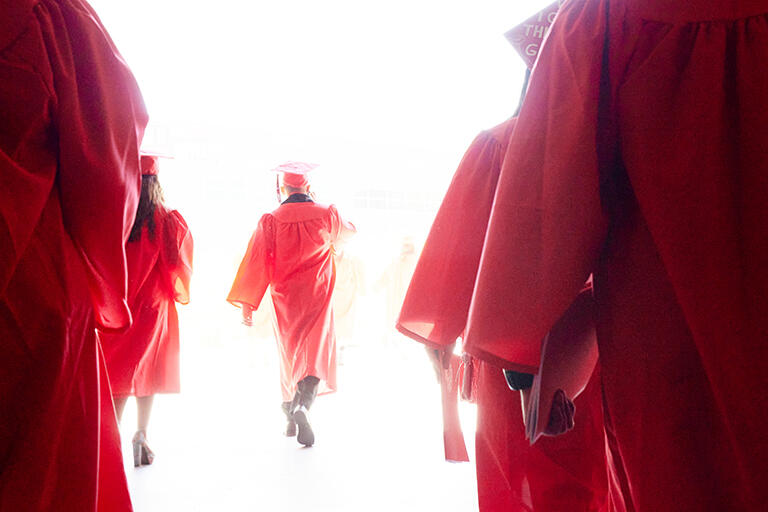 Back view of two lines of students wearing red commencement robes, one student is in the middle walking into a bright light
