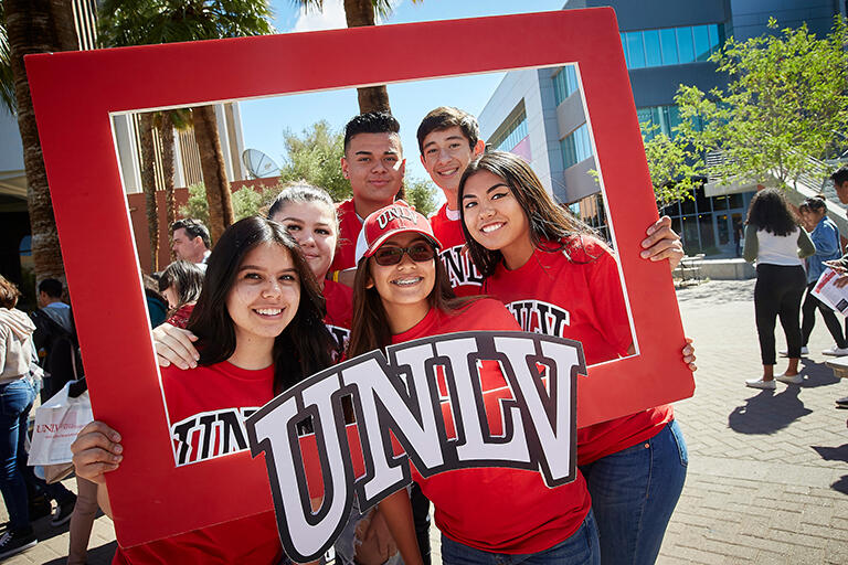 Un grupo de cinco estudiantes vestidos con camisetas rojas de UNLV. Están sosteniendo un cuadro rojo de UNLV por sus cabezas para imitar un cuadro de estilo polaroid.