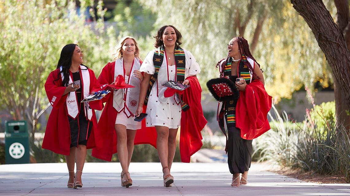 Four students walking on campus in cap and gown