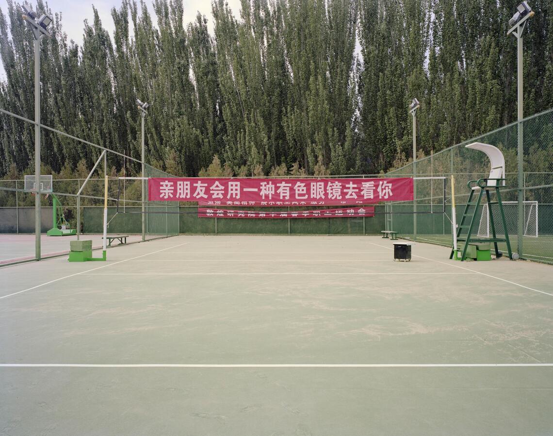 Long red banners hang on the nets of a public tennis court. White text written in Chinese characters is printed along the length of each banner. Tall green trees stand behind the courts.