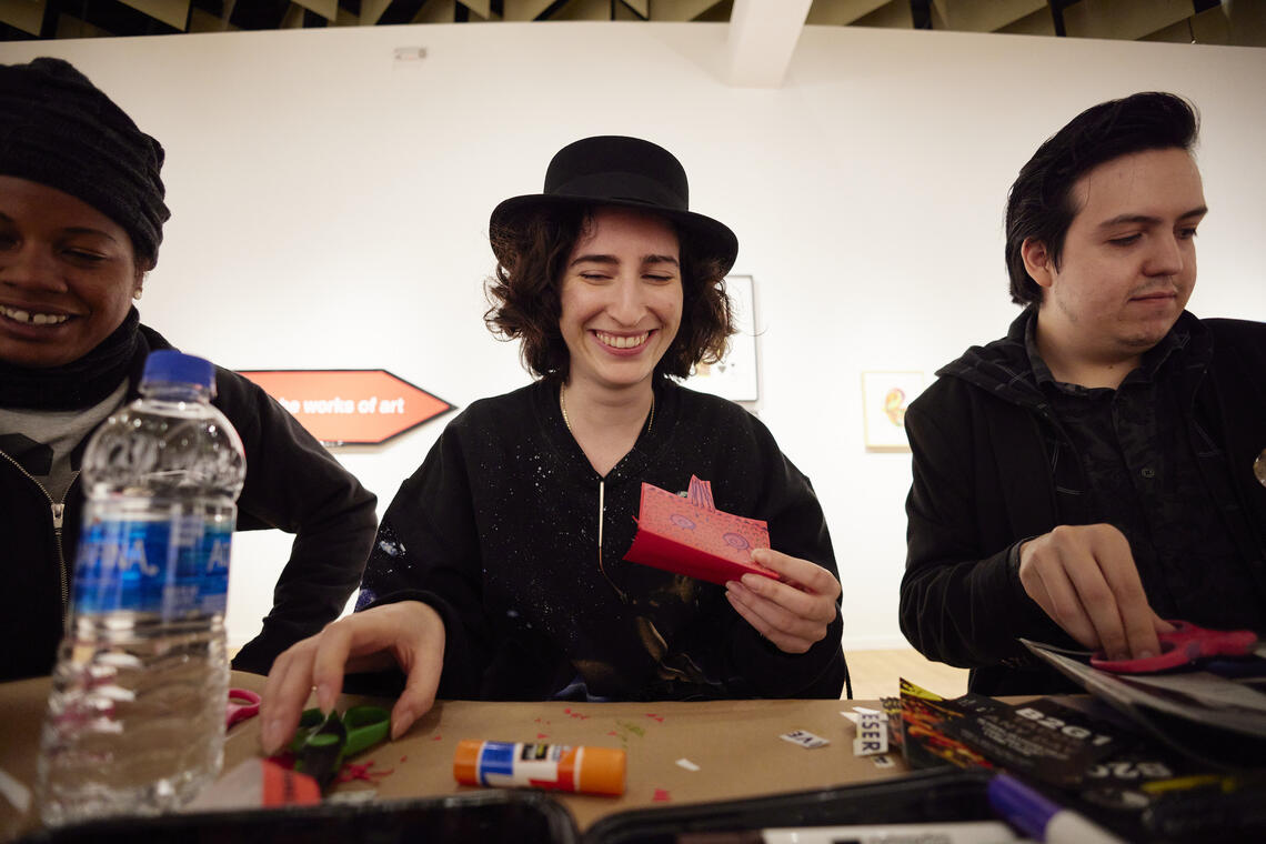 Three people sit at a table, looking happy as they make things with scissors, paper, and glue. The person in the middle is holding up a small folded paper zine and smiling broadly.