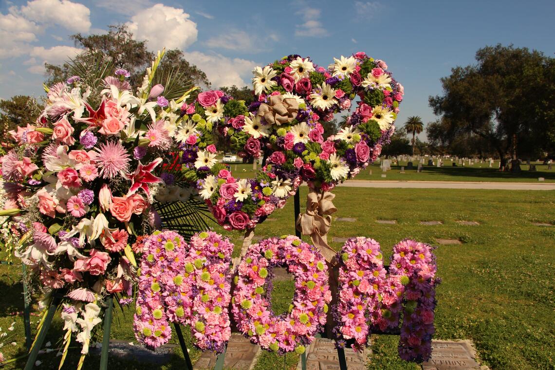 The word &quot;MOM,&quot; crafted out of pink flowers, stands next to a large arrangement of roses, daisies, irises, and other decorative blooms. Rows of grave markers are embedded in the sunlit lawn around them.
