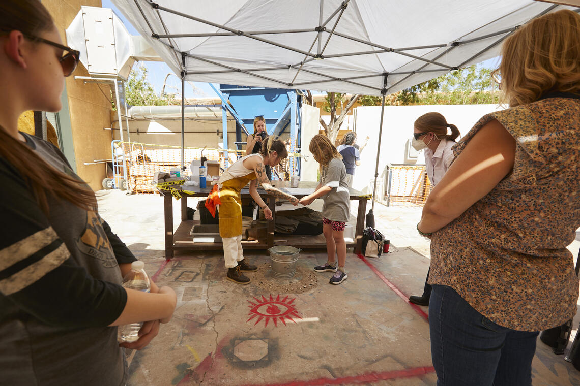 Several people watch as a woman wearing a leather apron helps a small girl hold a ladle over a water-filled metal bucket. A stylized red eye is printed on the concrete by their feet. Everyone in this picture is standing under an awning on a sunny day outdoors.