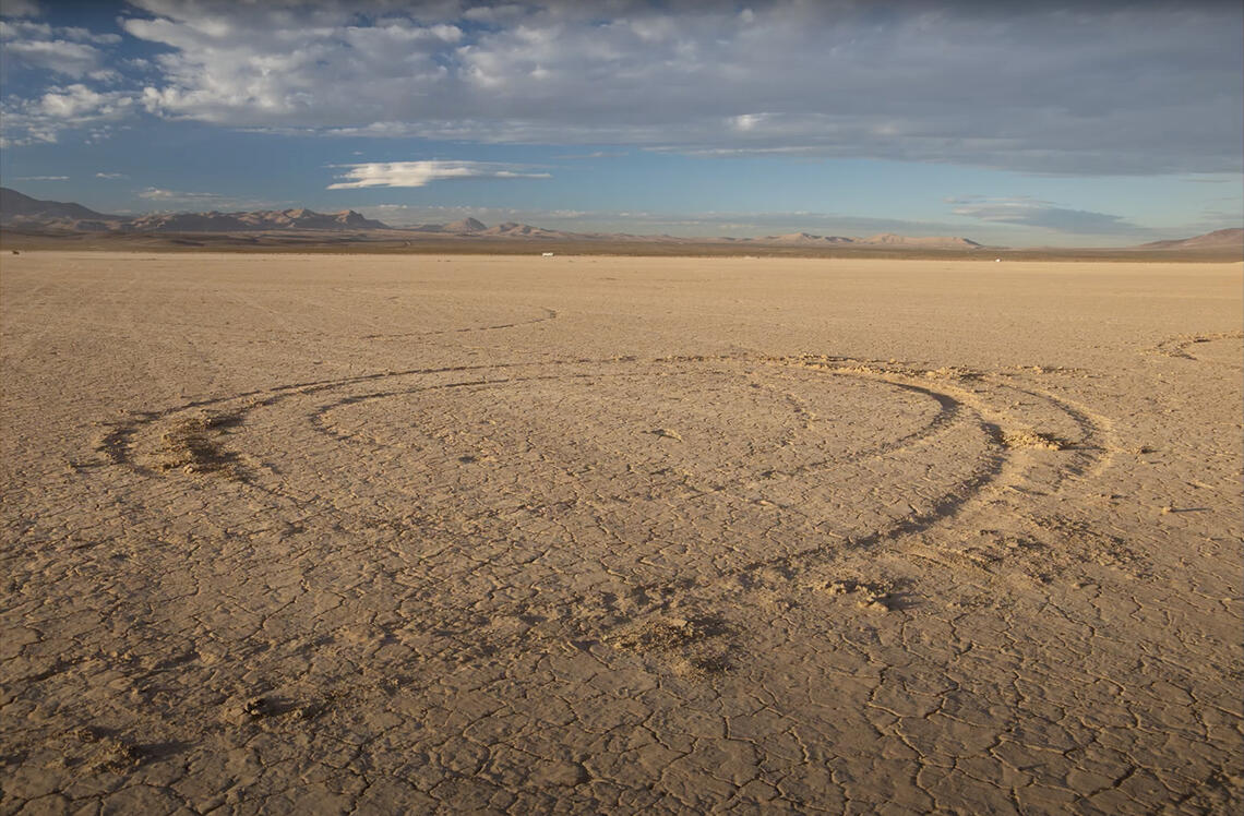 looking out across a vast flat desert plain we see a circle of wheel tracks created by a vehicle. The vehicle itself is absent.