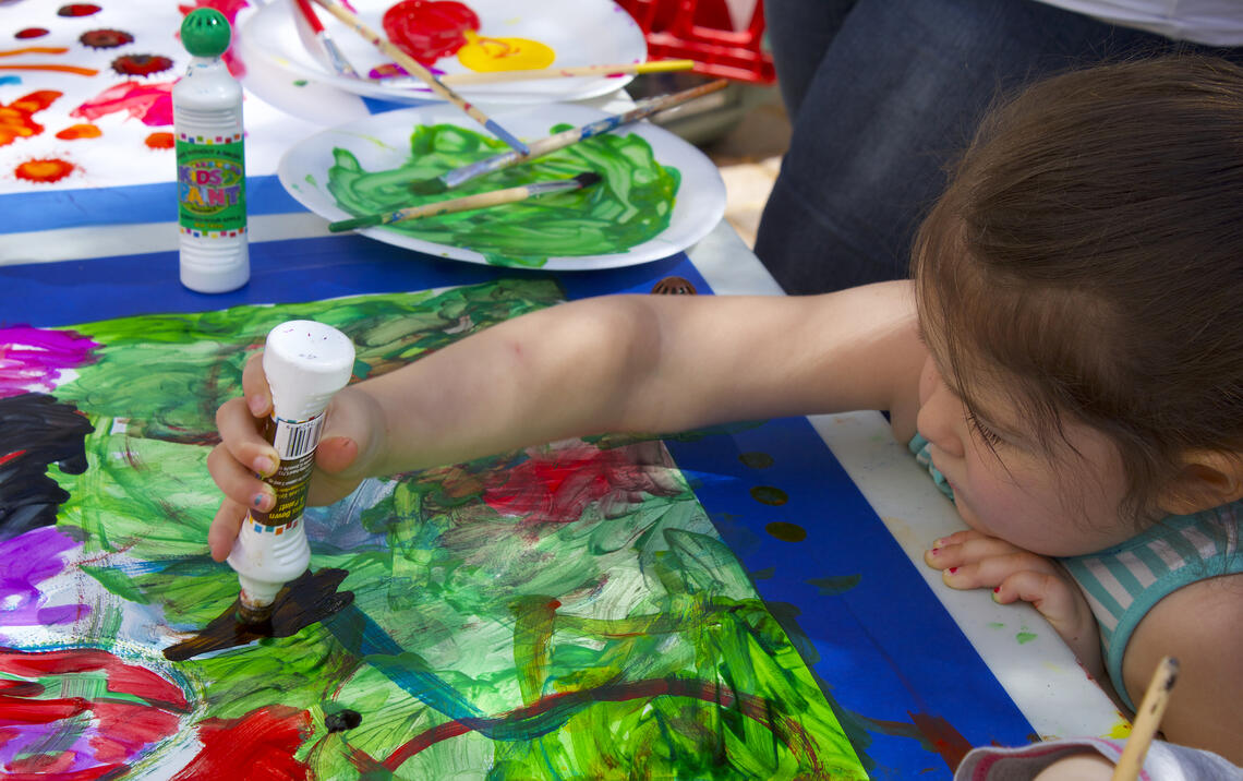 A small child stretches her arm over a table to add paint from a kid-sized paint dauber to a sheet of paper that is already covered with paint.