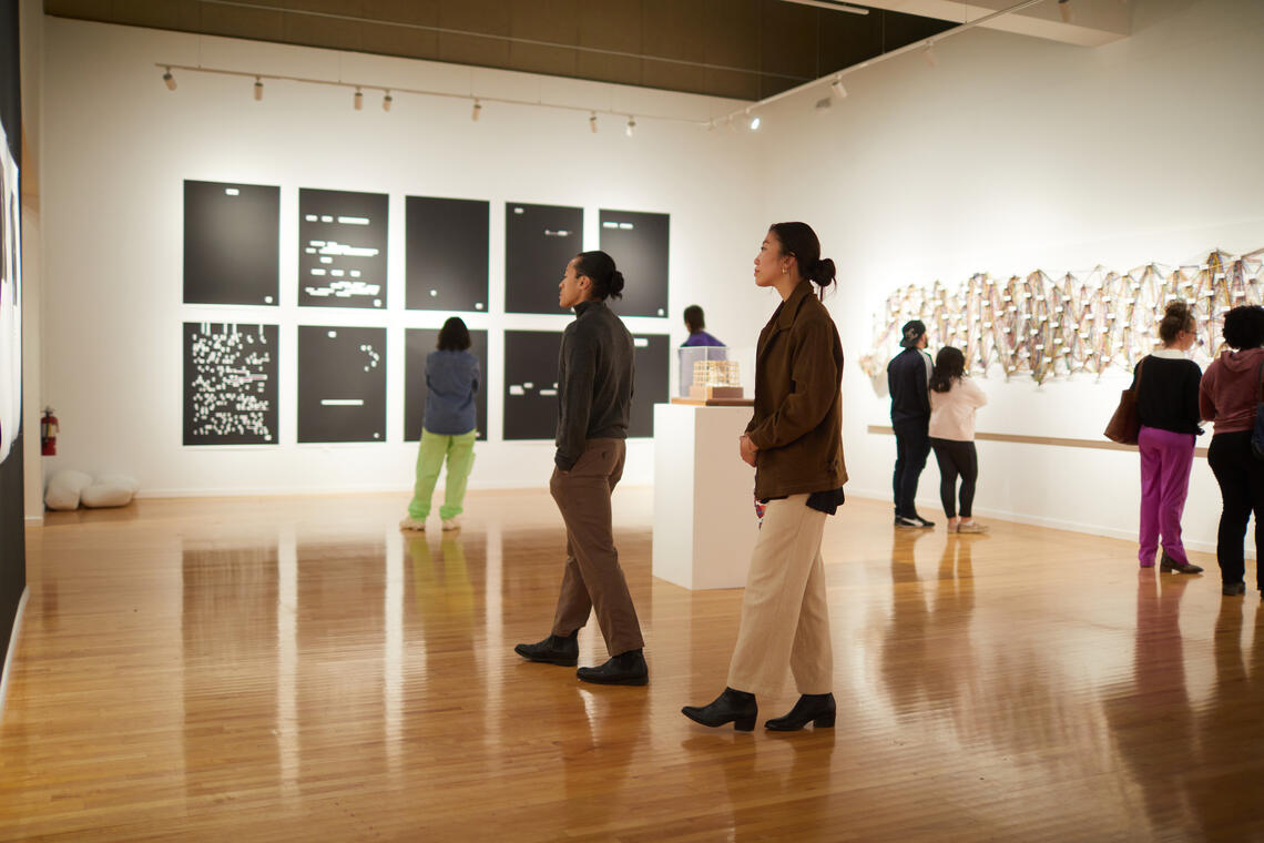 Neatly-dressed people are walking around a large, well-lit room with white walls and a wooden floor. They are paying attention to artwork on the walls. Rows of dark panels hang on the farthest wall. A long multi-colored artwork hangs on an adjacent wall. The artworks are too far away to reveal details.
