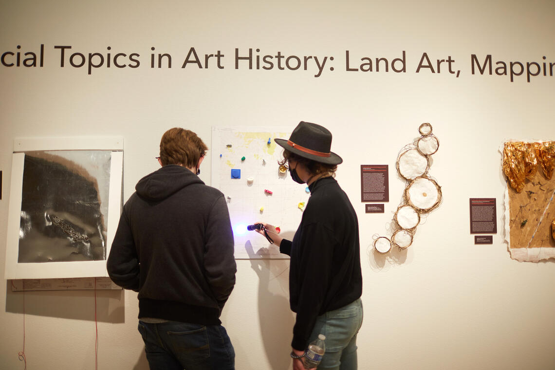 Two people stand in front of a wall of artworks with their backs to the camera. One of them is shining a flashlight onto the piece in front of them: a square map decorated with small three-dimensional objects in different colors. Text on the wall above them reads: “… cial Topics in Art History: Land Art, Mappi …”