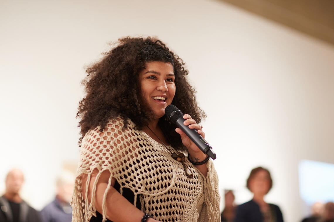 A Black woman with stands in a high-ceilinged room speaking into a microphone. She is dressed in black, with a shawl thrown over her shoulders. A crowd is standing around her - we can see their heads in the background.