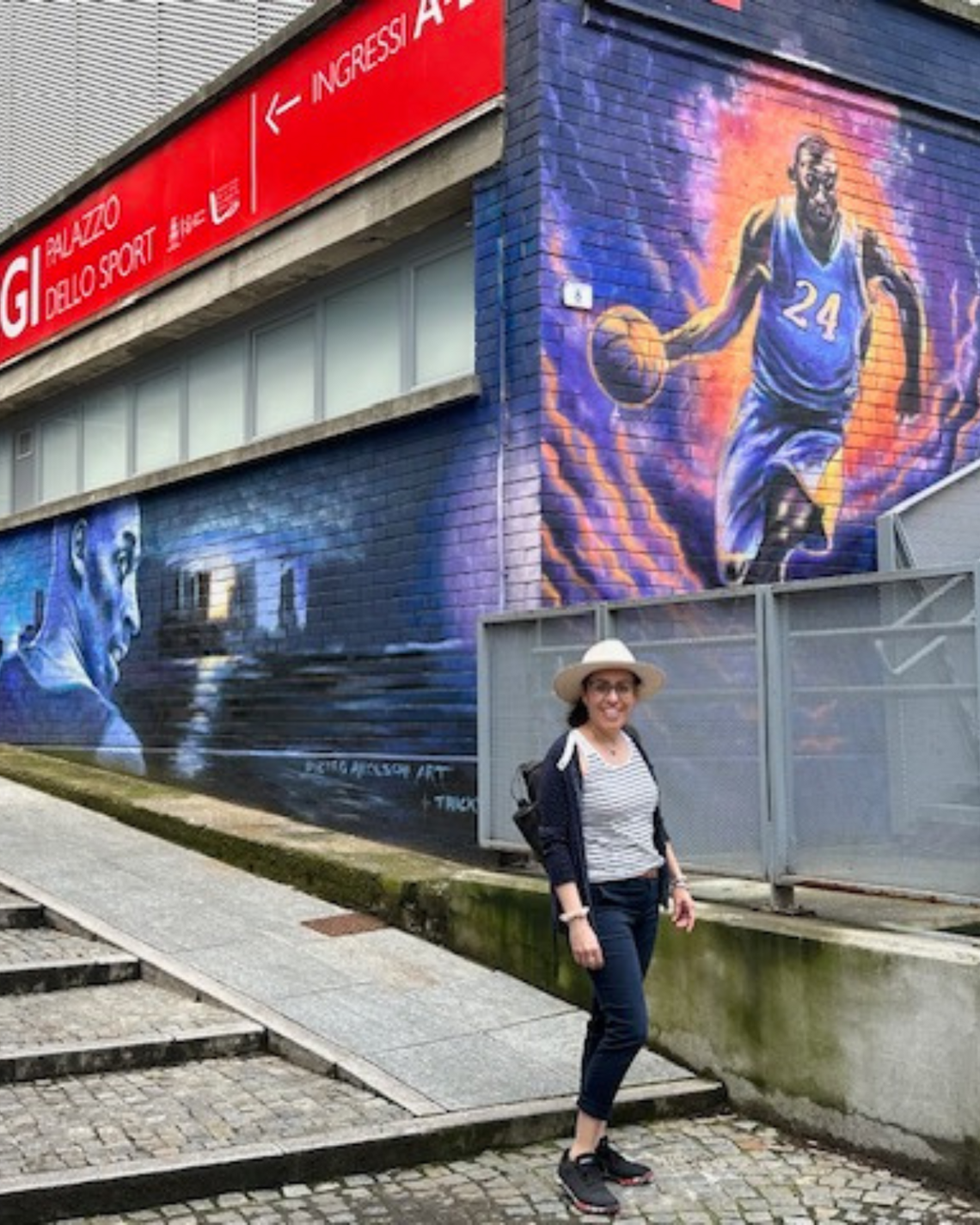 A woman in a hat posing in front of a Kobe Bryant mural in Italy.