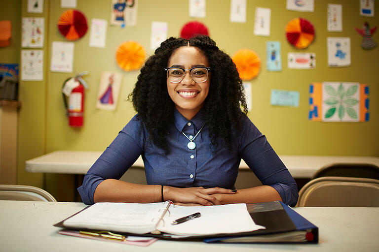 A woman smiling sitting down at a table with a notebook and a pencil in a classroom