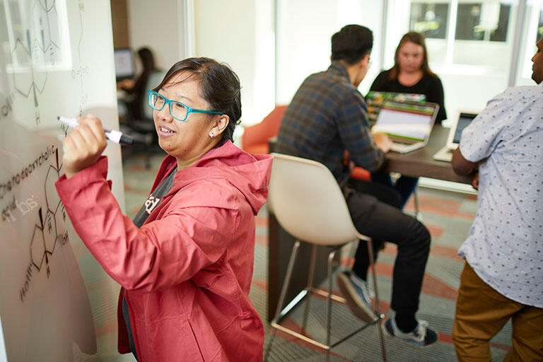 A woman wearing a pink jacket and blue glasses writing on a whiteboard