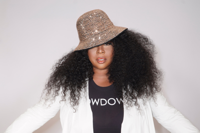 Picture of Ms. Monét standing wearing a white jacket, black shirt and tan color hat.