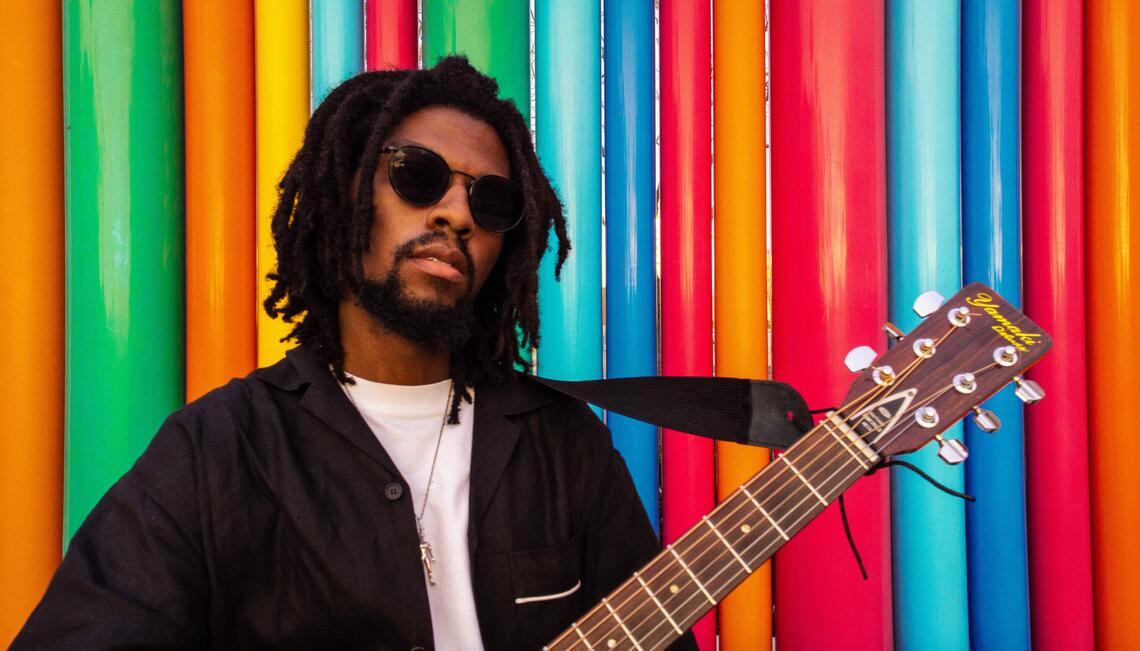 A man with dreadlocks and sunglasses holds a guitar in a dynamic position as if he is about to play. His black jacket, guitar strap, and hair stand out vividly against a bright, striped wall made from vertical pipes painted in different colors.