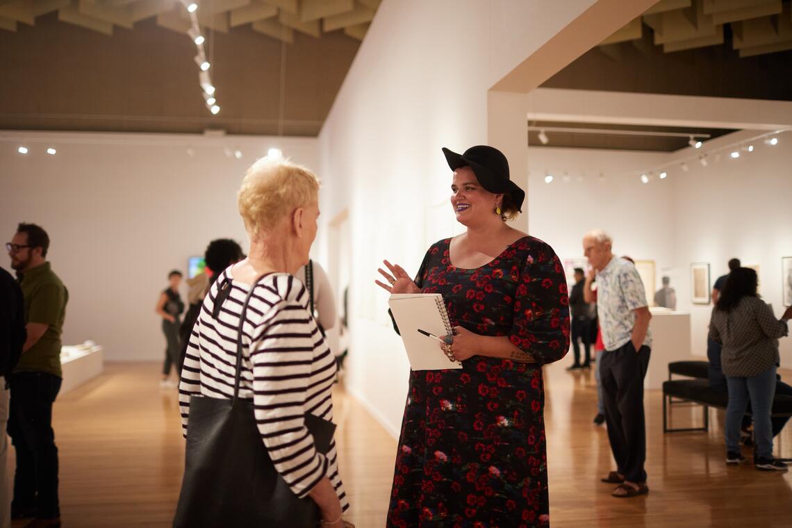 Two women stand in a big bright gallery space with white walls and a high ceiling. Groups of people are looking around the room curiously behind them. The taller one is saying something with an effusive, excited expression as she holds up a blank notebook.