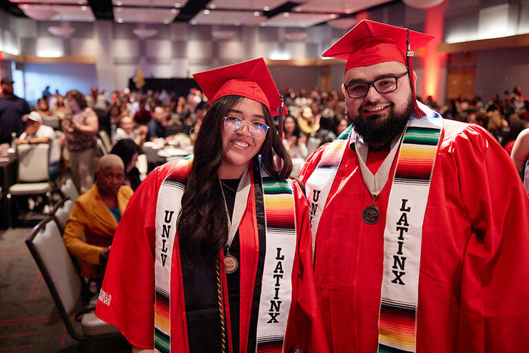 Two latinx graduates wearing red commencement caps and gowns and the U.N.L.V. latinx sashes
