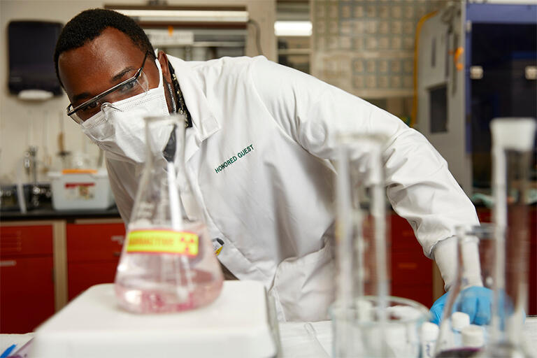 guy wearing lab coat, safety goggles, and mask studying a beaker with chemicals inside