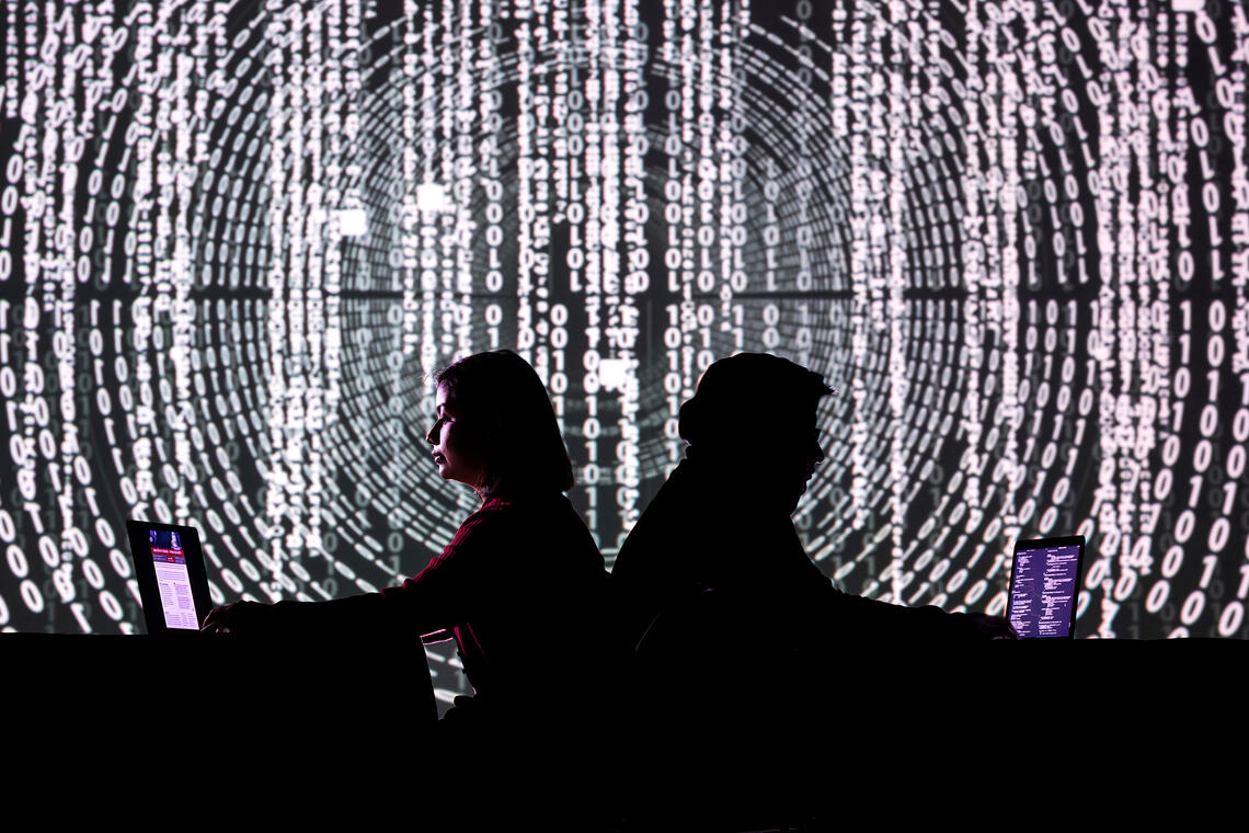 Two people sitting back to back, looking at their laptops with Binary code projecting on the wall