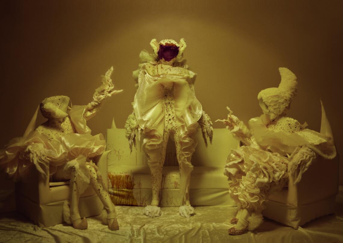 A profoundly yellow space with two yellow vinyl chairs on either side of a yellow couch. Two costumed yellow figures sit on the chairs, reacting in shock to a third yellow figure who stands between them. Al three figures are costumed like beautiful monsters, with extravagant ruffled torsos, pointed feet, and long fluffy fingers. The face of the standing figure, however, is red with blood. More blood is spattered over the couch.