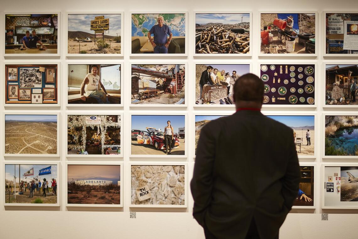 A man in a dark suit stands with his back to us. He is looking at rows of framed photographs hanging on the wall. The photos depict people and landscapes from a desert environment--sandy countryside scattered with small bushes; a prospector tipping water into a bucket; nuggets in containers, a rally in the sand.