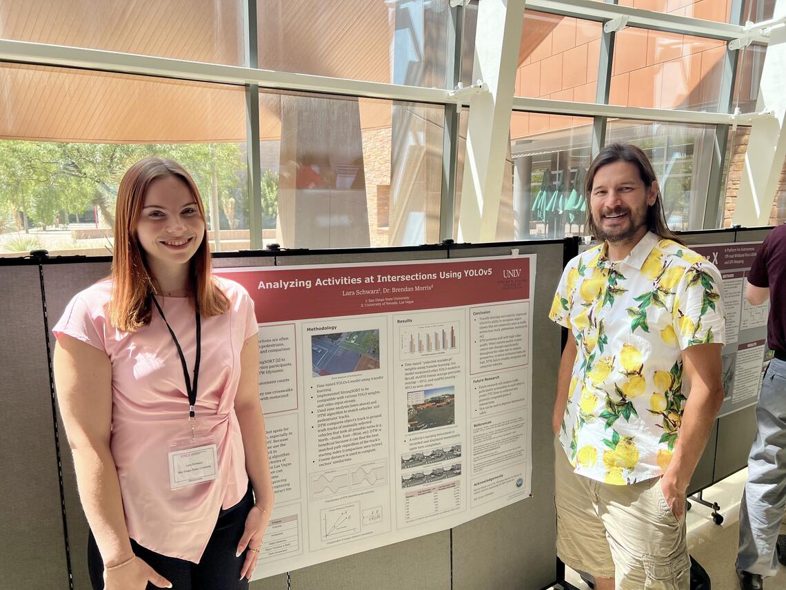 Two people pose for a photo in front of a research poster with a bank of floor-to-ceiling windows behind them