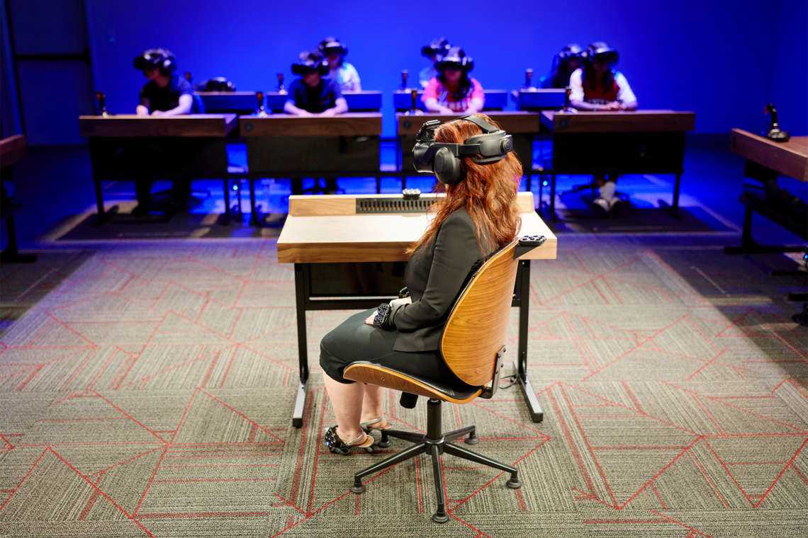 Seven people sitting at individual desks with virtual reality headsets as someone sits center of the room by a desk with a headset