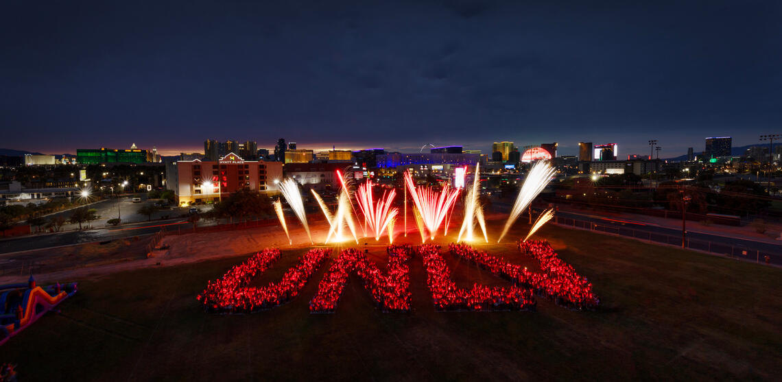 aerial shot of humans spelling out UNLV with fireworks behind the letters
