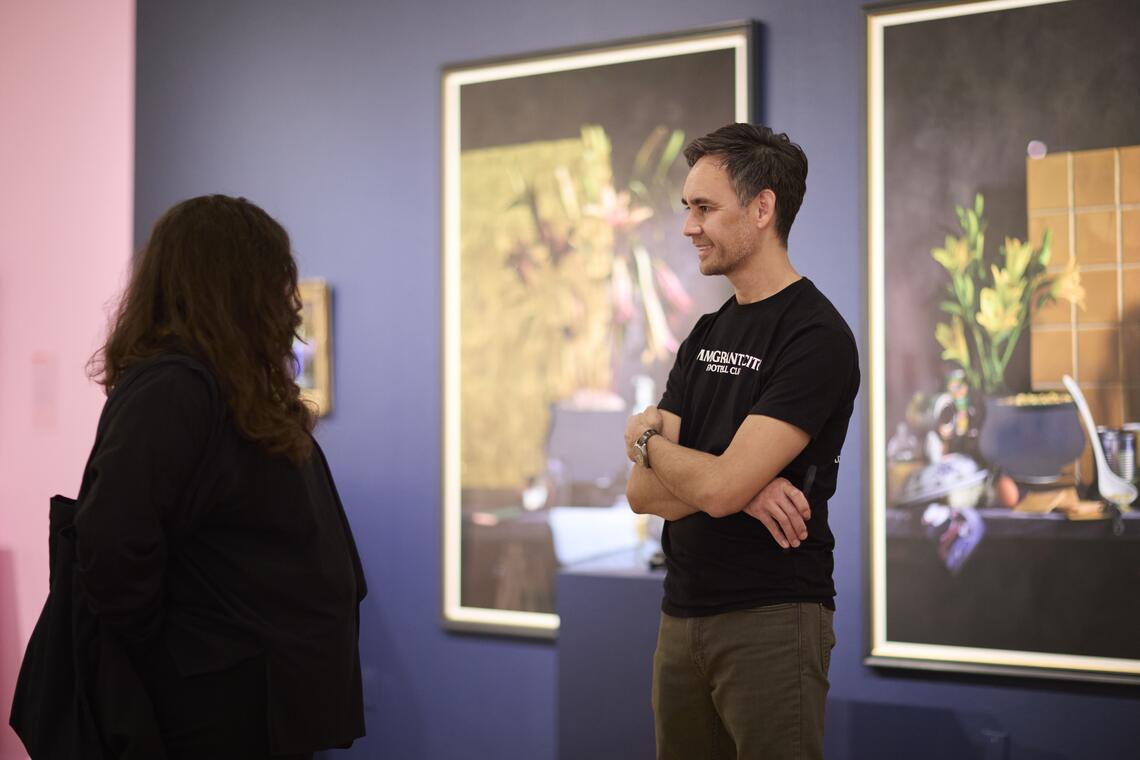 A man in a casual black t-shirt smiles as he talk to a woman with long dark hair. Large framed still life images of flowers, pots, and spoons hang on the wall behind him.
